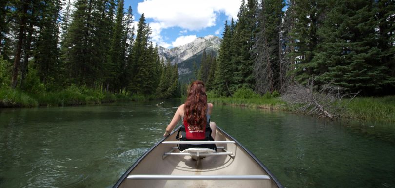 Woman canoeing on a calm river surrounded by evergreen trees and mountains