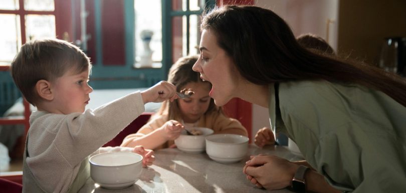 Young child feeding a parent during a family meal while another child eats at the table